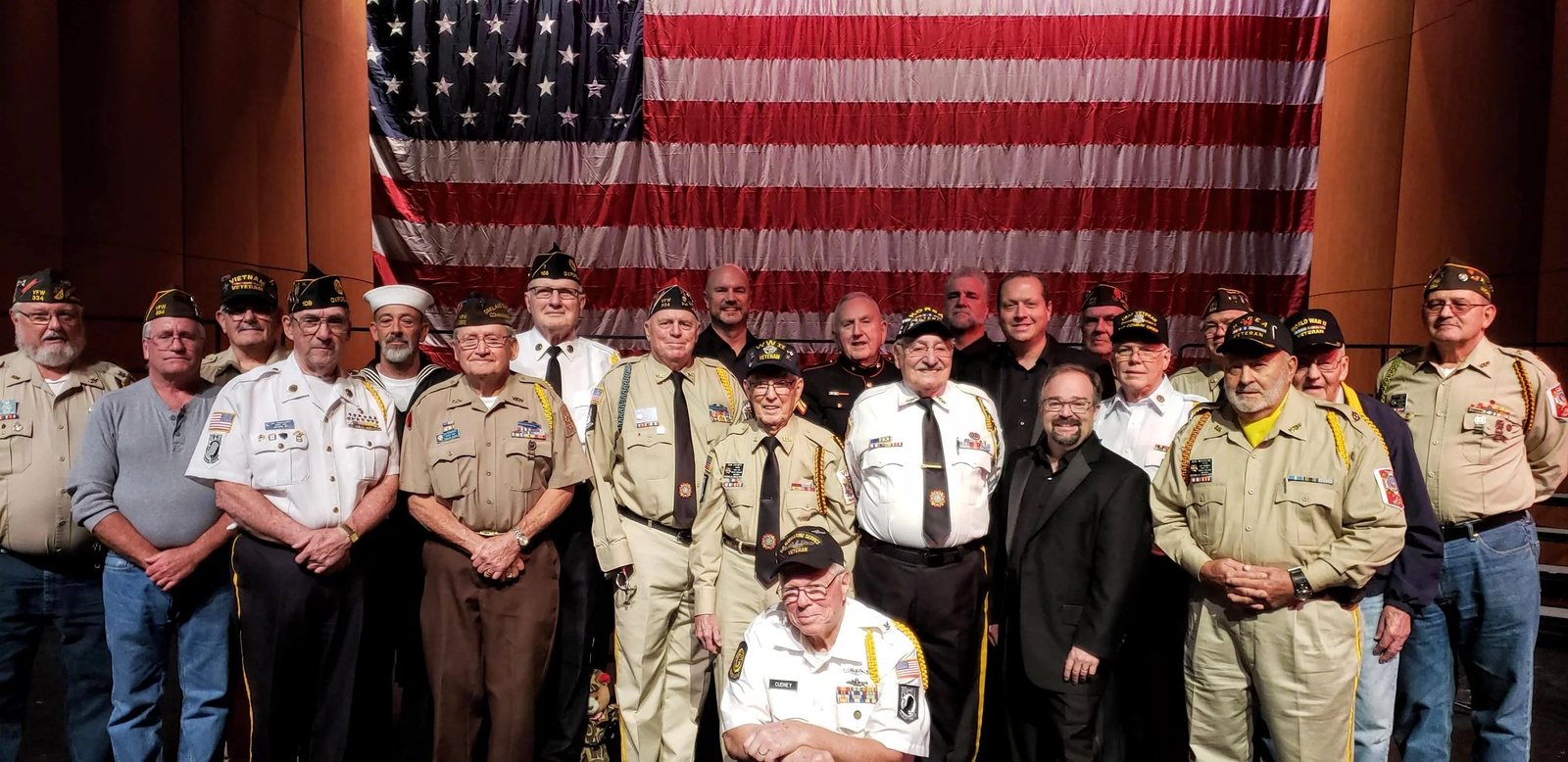Three Men and a Tenor standing among more than twenty veterans on stage in Ypsilanti, Michigan, in front of a large American flag. Greg Cudney, in Navy dress whites, is seated at the front.